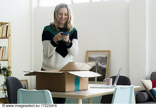 Smiling woman taking photo of package box through mobile phone at home