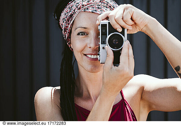 Smiling woman taking photo through camera while standing against wall