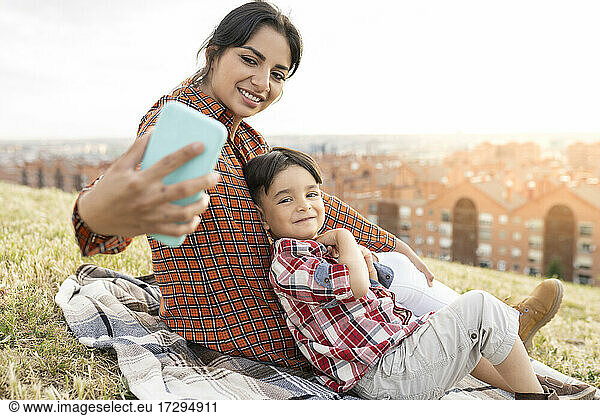 Smiling woman taking selfie with boy while sitting on hill