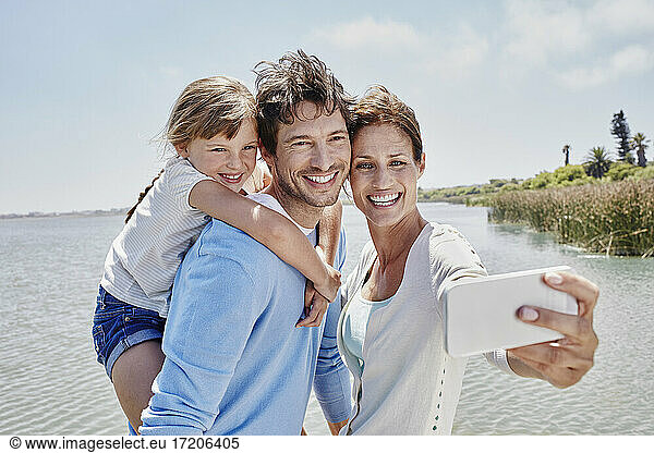 Smiling woman taking selfie with family by lake on sunny day