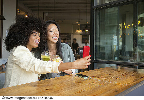 Smiling woman taking selfie with friend holding healthy drink at cafe