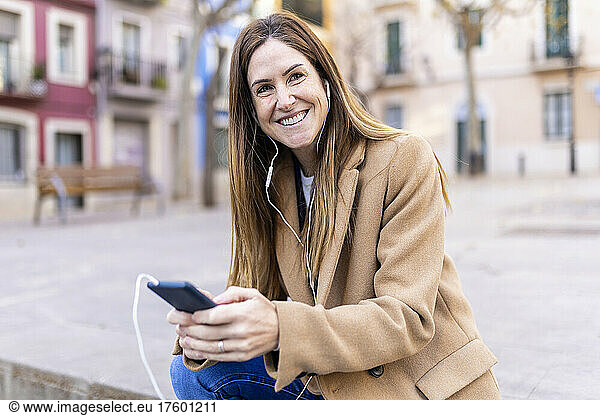 Smiling woman talking on mobile phone through in-ear headphones at city street
