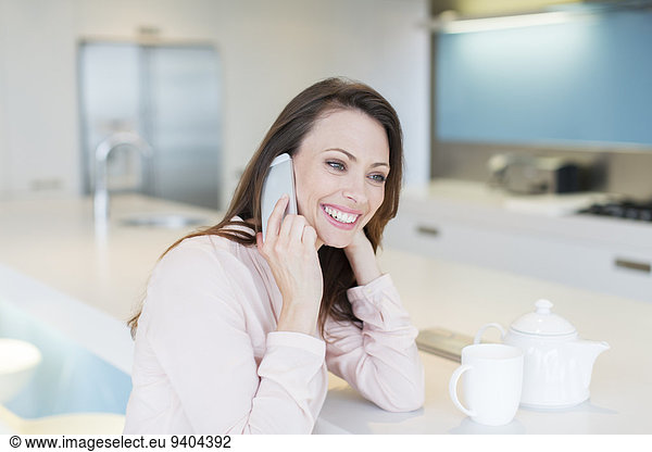 Smiling woman talking on phone in kitchen  tea cup and tea pot on counter