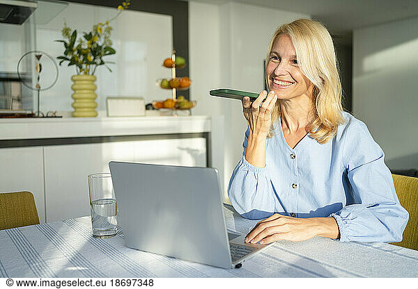 Smiling woman talking on speaker phone with laptop at desk in home office