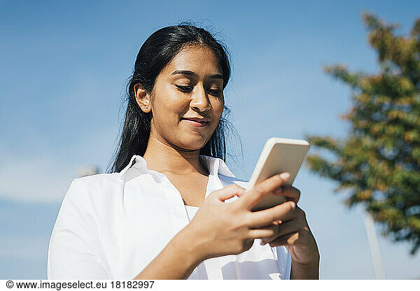 Smiling woman text messaging through mobile phone under blue sky