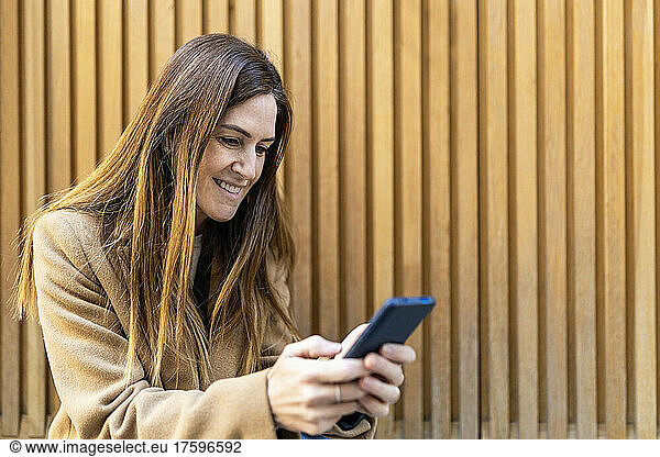 Smiling woman text messaging through smart phone in front of wall