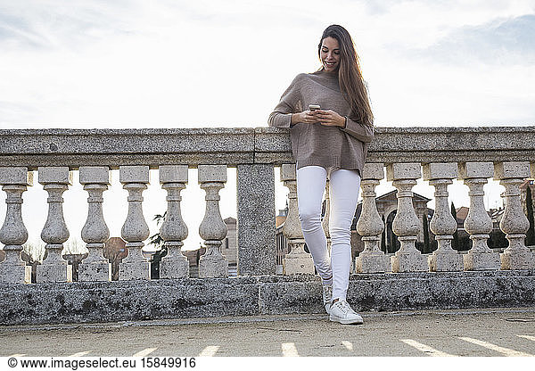 Smiling woman using her phone while leaning on stone railing