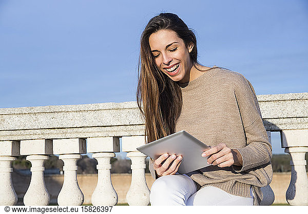 Smiling woman using her tablet while sitting on a stone bench