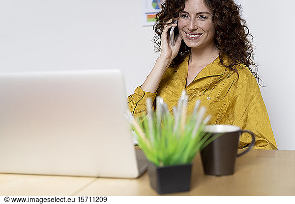 Smiling woman using laptop and smartphone at desk in office