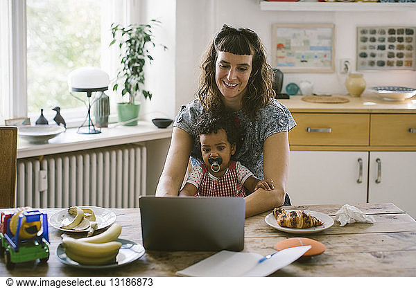 Smiling woman using laptop while sitting with daughter at dining table in house