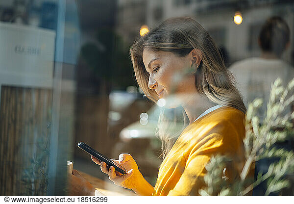 Smiling woman using smart phone in cafe seen through glass