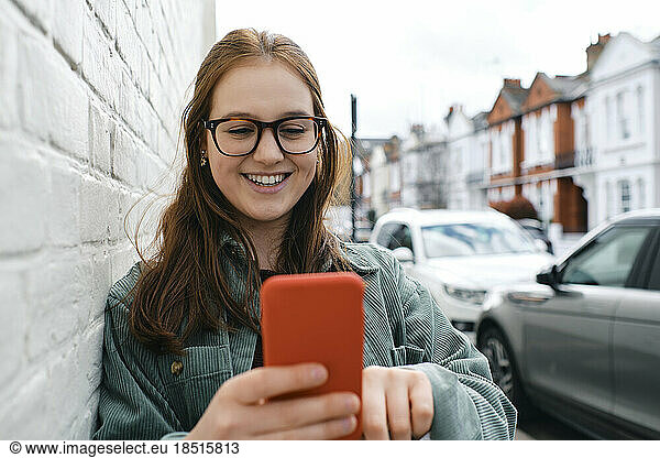 Smiling woman using smart phone leaning on wall