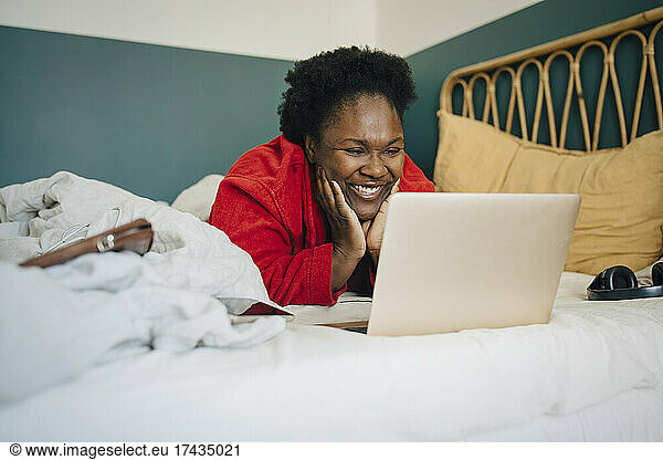 Smiling woman watching on laptop while lying on bed at home