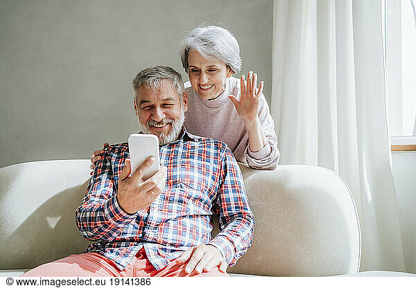 Smiling woman waving with man video calling through smart phone at home