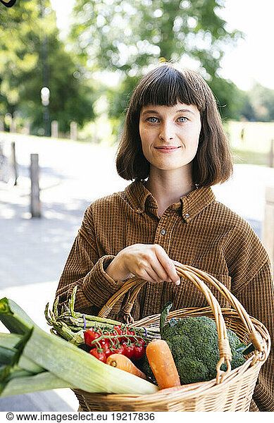 Smiling woman wearing brown shirt holding basket of vegetables at farmer's market