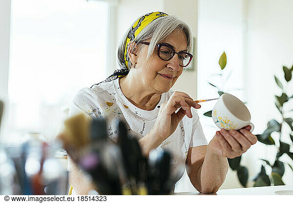 Smiling woman wearing eyeglasses painting ceramic cup in workshop