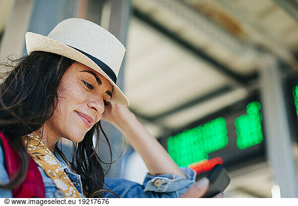 Smiling woman wearing hat using mobile phone at tram station