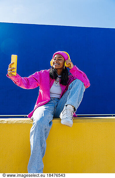 Smiling woman wearing headphones and taking selfie through mobile phone in front of blue wall