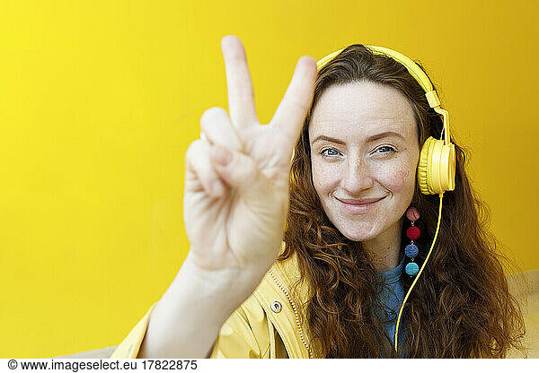 Smiling woman wearing headphones gesturing peace sign in front of yellow wall