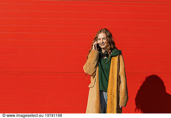 Smiling woman wearing jacket talking on smart phone in front of red wall