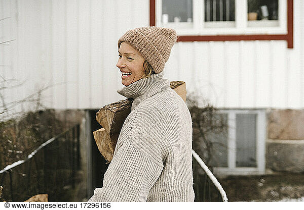 Smiling woman wearing knit hat carrying firewood while looking away