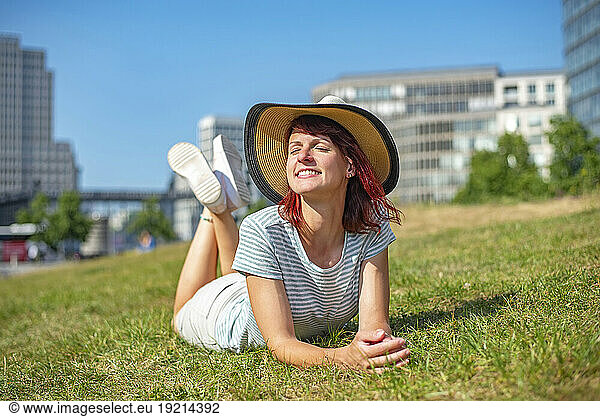 Smiling woman wearing sun hat relaxing on grass with eyes closed