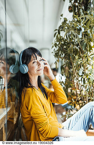 Smiling woman wearing wireless headphones listening to music by glass
