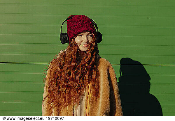 Smiling woman wearing wireless headphones listening to music in front of green wall