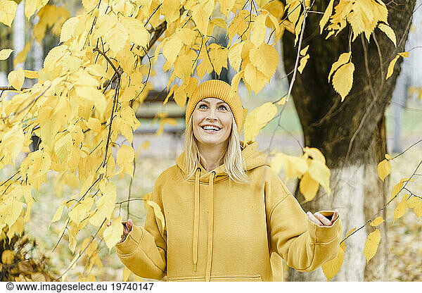Smiling woman wearing yellow hooded shirt and touching leaves of tree in park