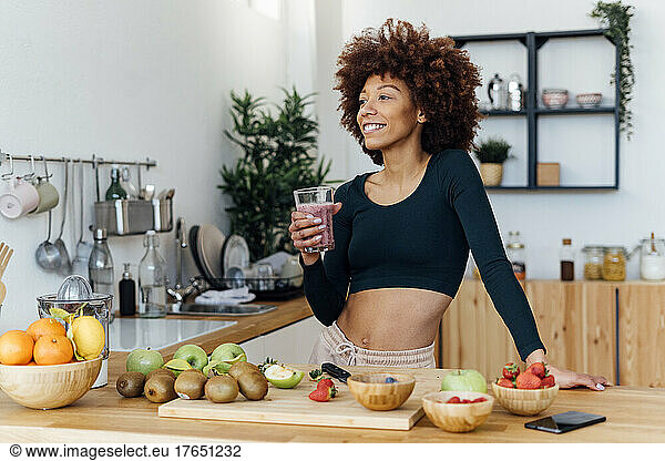 Smiling woman with Afro hairstyle holding glass of smoothie standing in kitchen