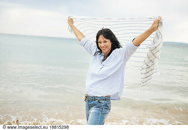 Smiling woman with arms raised holding scarf at beach