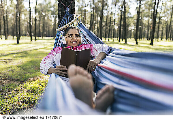 Smiling woman with book lying in hammock listening music at park