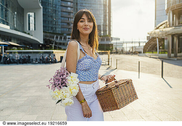 Smiling woman with bouquet of flowers and basket standing on footpath