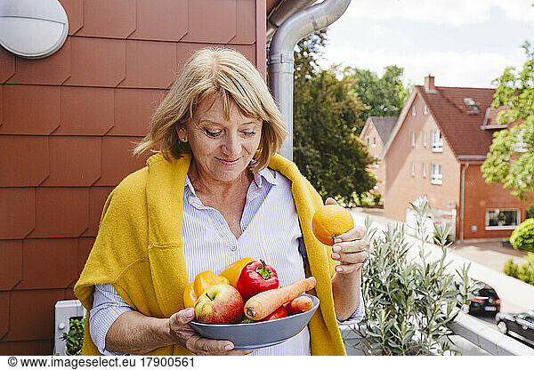Smiling woman with bowl of fresh fruits and vegetables standing in balcony