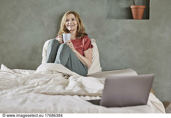 Smiling woman with coffee cup sitting on bed at home