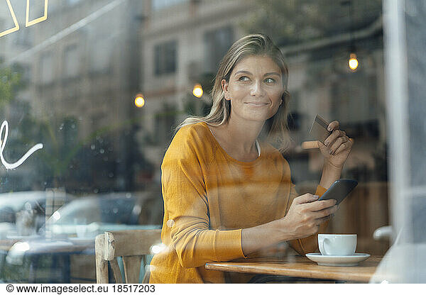 Smiling woman with credit card and smart phone sitting in cafe