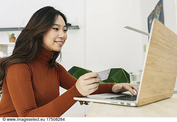 Smiling woman with credit card paying through laptop at home