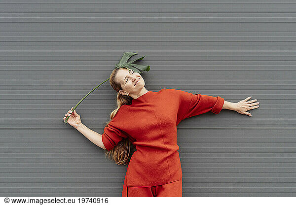 Smiling woman with eyes closed holding monstera leaf in front of gray wall