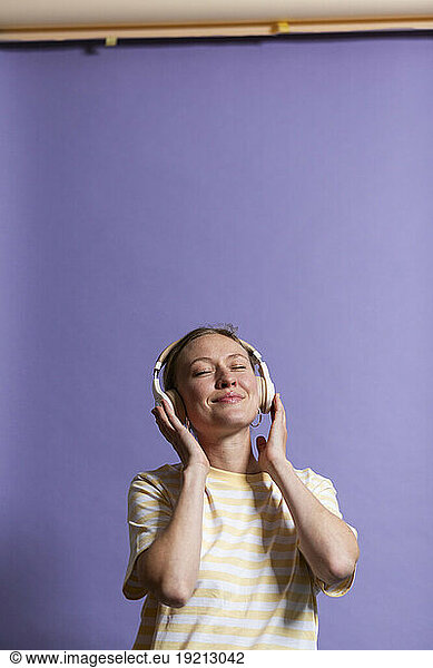 Smiling woman with eyes closed listening music on headphones against purple background