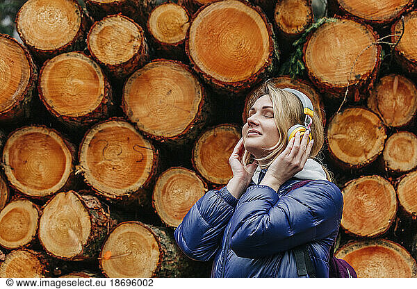 Smiling woman with eyes closed listening music through headphones by stack of logs