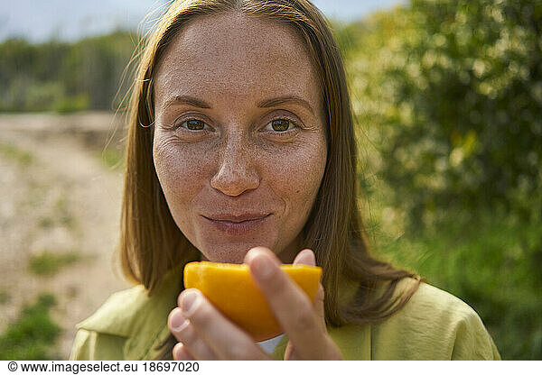 Smiling woman with freckles on face holding orange slice in orchard