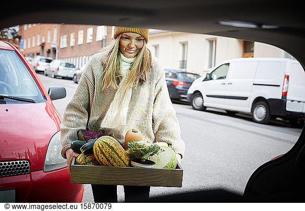 Smiling woman with freshly produce vegetables in container standing near car trunk