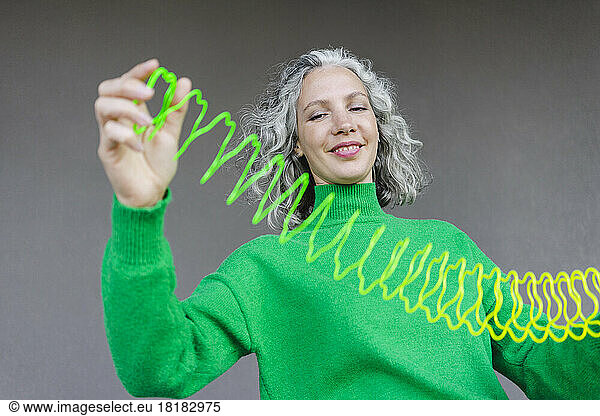 Smiling woman with gray hair stretching metal coil toy in front of wall