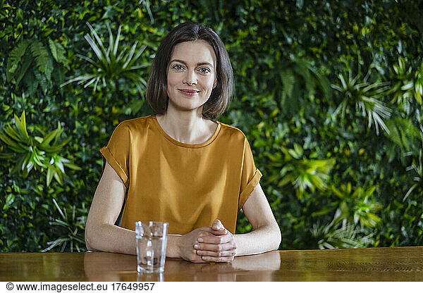 Smiling woman with hands clasped sitting by table at wall with plants