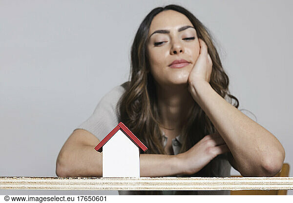 Smiling woman with head in hand looking at model of house against gray background