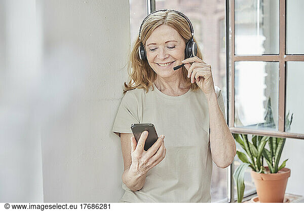 Smiling woman with headset using smart phone standing in front of window at home