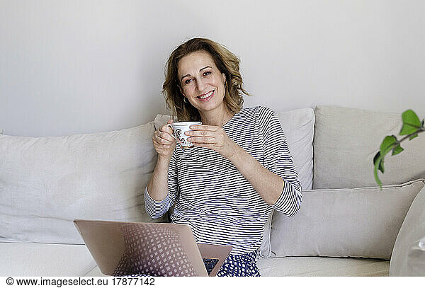 Smiling woman with laptop and coffee cup sitting on couch at home