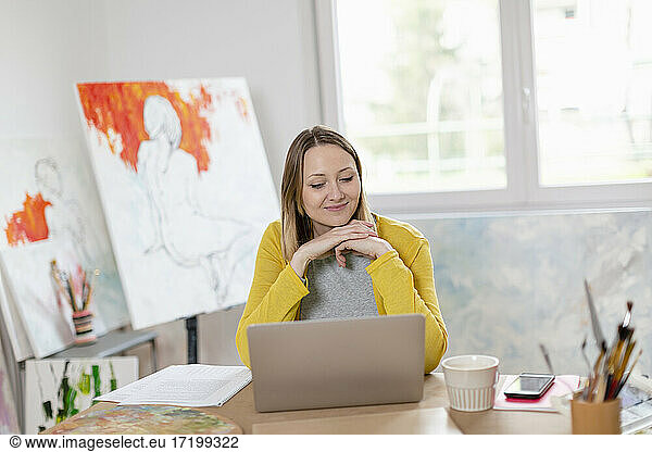 Smiling woman with laptop on table while sitting at home studio