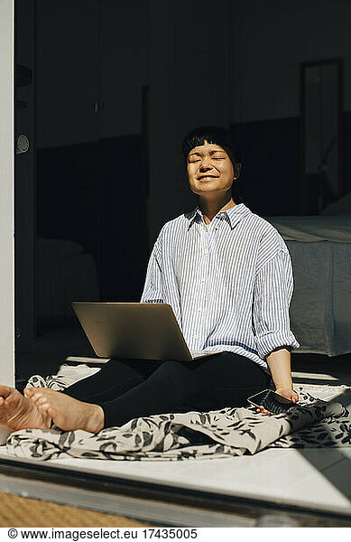 Smiling woman with laptop sitting in sunlight at home