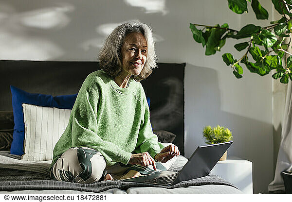 Smiling woman with laptop sitting on bed at home